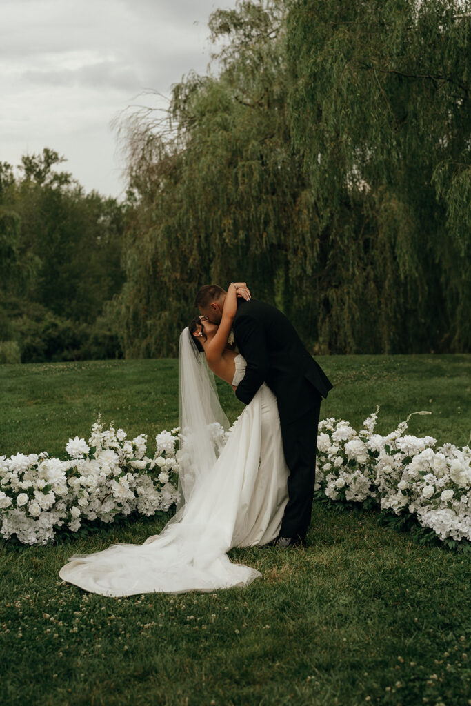 Rhode Island Wedding Photographer | Aisha Lee Photography Cinematic Wedding Photographer | A bride and groom embrace in a tender kiss outdoors, surrounded by lush greenery and white flowers. The bride wears a flowing gown, adding a romantic feel.