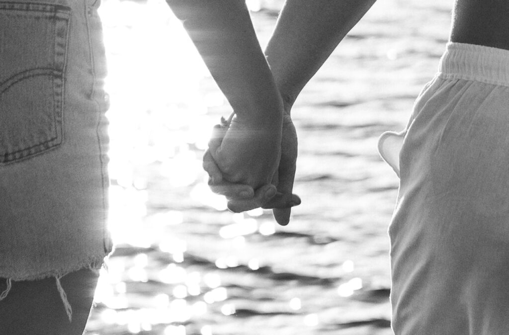 Oceanfront Weddings New England | Aisha Lee Photography Cinematic Wedding Photographer | A couple holding hands in front of shimmering water. The black and white image conveys a serene and intimate moment, with sunlight reflecting on the waves.