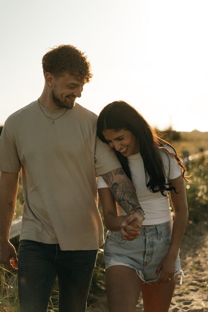 Adventure Wedding and Elopement Photographer New England | Aisha Lee Photography Cinematic Wedding Photographer | A couple walks hand in hand on a sunny day, smiling warmly. The woman, in a white top and denim shorts, leans affectionately on the man in a beige shirt.
