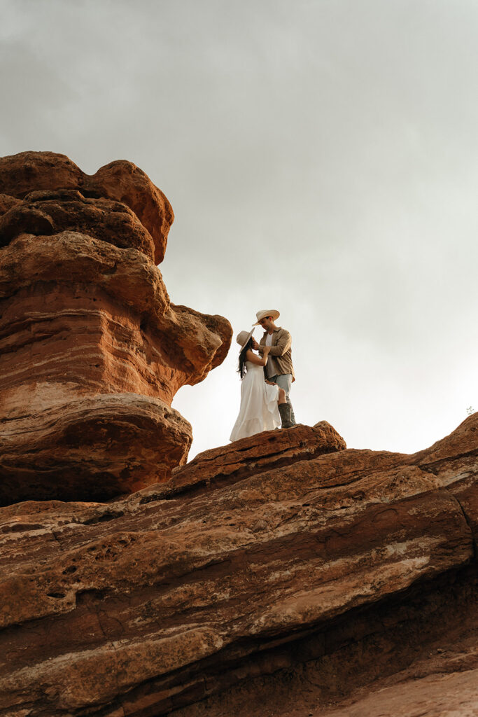 Destination Engagement Photographer New England | Aisha Lee Photography Cinematic Wedding Photographer | A couple stands together on a rocky ledge under a cloudy sky. The woman wears a white dress and the man wears a hat and jacket.