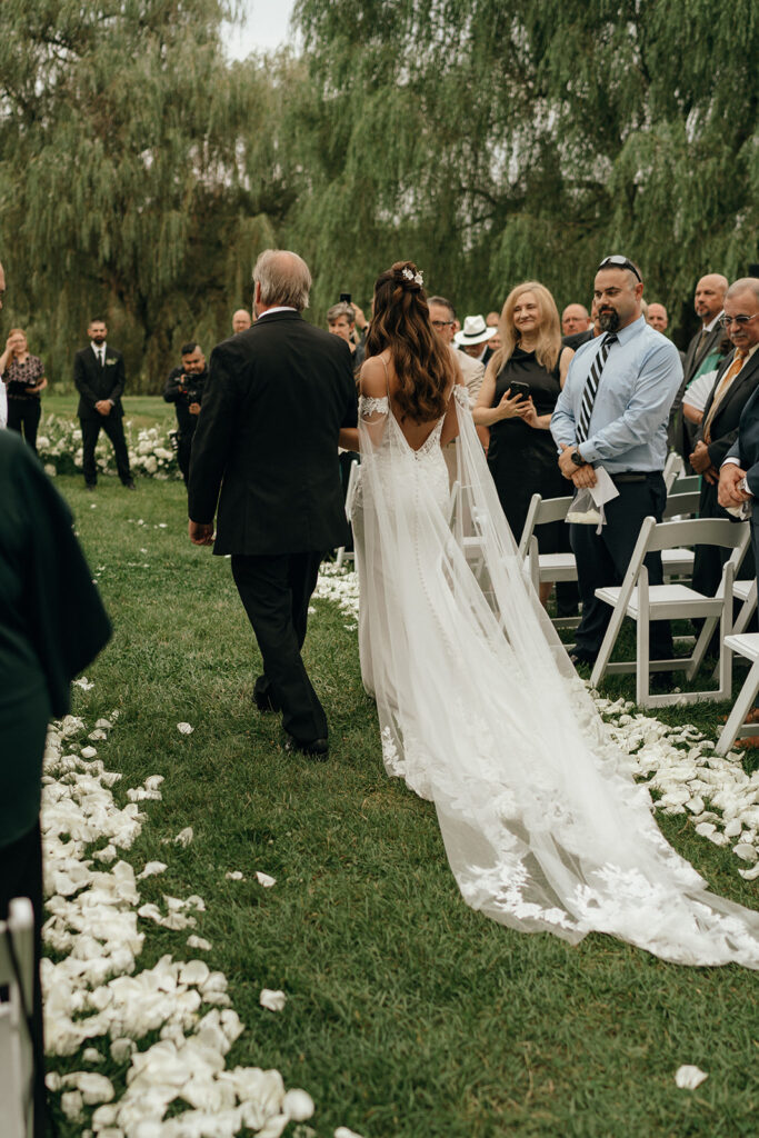 New England Beach Wedding | Aisha Lee Photography Cinematic Wedding Photographer | Bride in an elegant white gown walks down a petal-strewn aisle outside, escorted by a man. Guests stand on either side, creating a joyful atmosphere.