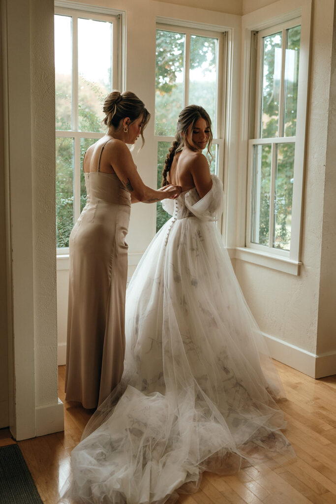 Unique Wedding Venues New England | Aisha Lee Photography Cinematic Wedding Photographer | A bride in an elegant white gown is being helped by a woman in a beige dress in a bright, airy room. The mood is joyful and intimate.