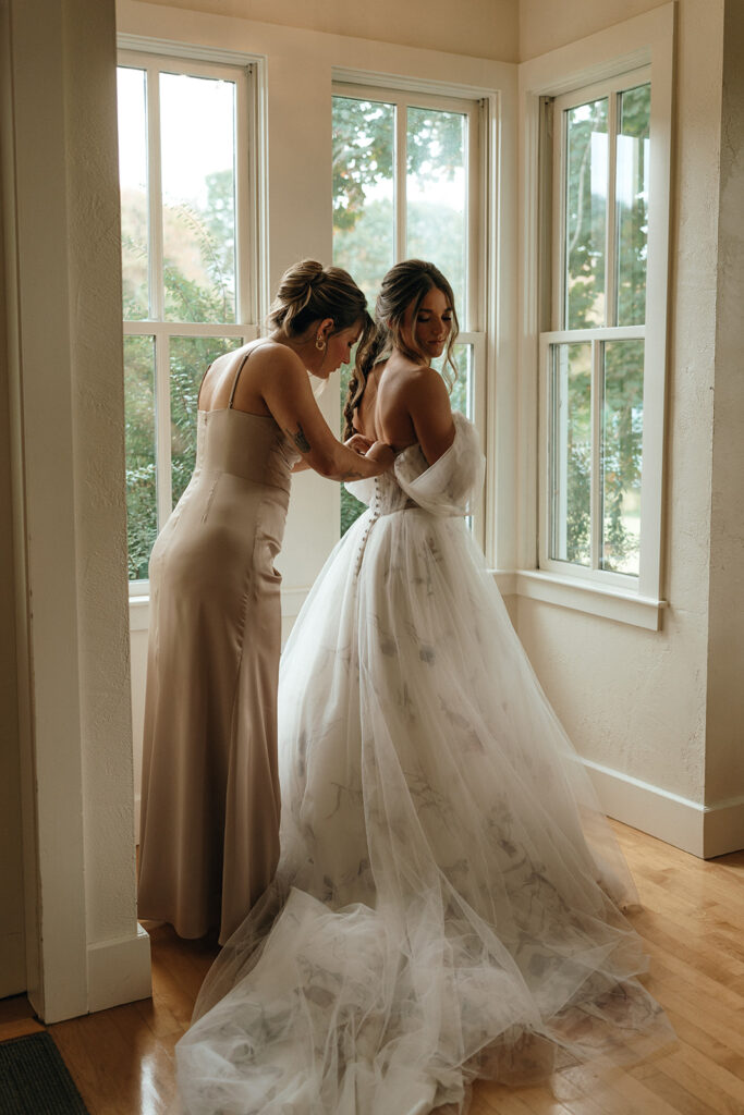 Castles in New England | Aisha Lee Photography Cinematic Wedding Photographer | Bride in a flowing white gown stands near large windows, lit by soft natural light. A woman in a beige dress helps fasten the gown, creating a serene, intimate moment.