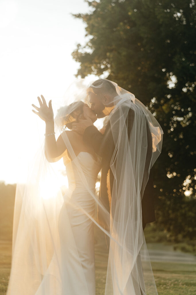 Eloping in New England | Aisha Lee Photography Cinematic Wedding Photographer | A couple kisses romantically under a long veil in front of a sunset. The warm light creates a dreamy atmosphere, symbolizing love and joy.