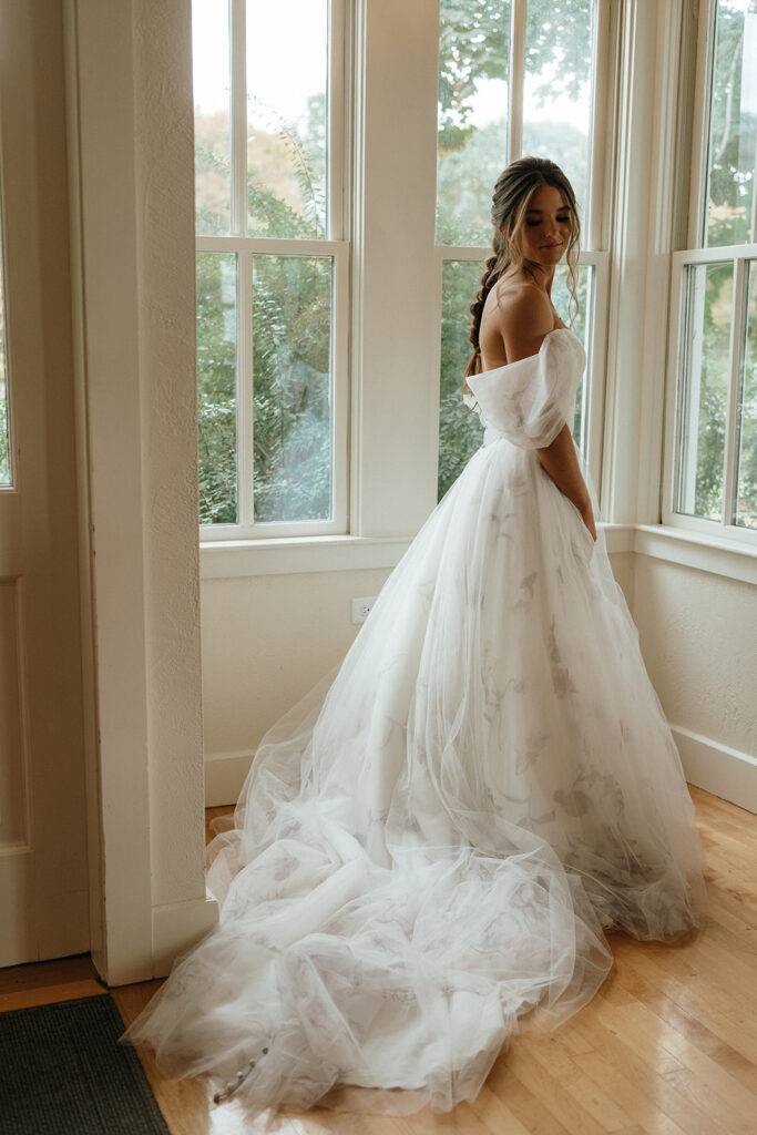 Top Wedding Venues in New England | Aisha Lee Photography Cinematic Wedding Photographer | A bride in an off-shoulder, tulle gown stands gracefully by large windows, with soft sunlight illuminating her and the room. The scene feels serene and joyful.
