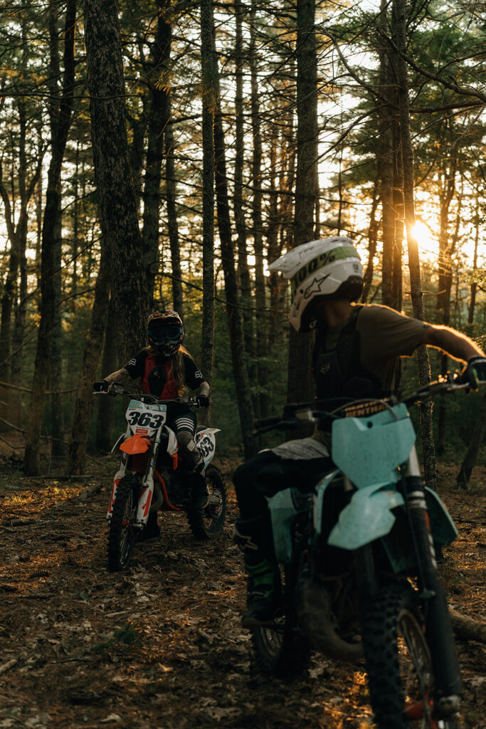 Adventure Wedding and Elopement Photographer New England | Aisha Lee Photography Cinematic Wedding Photographer | Two motocross riders navigate dirt bikes through a sunlit forest, casting long shadows.