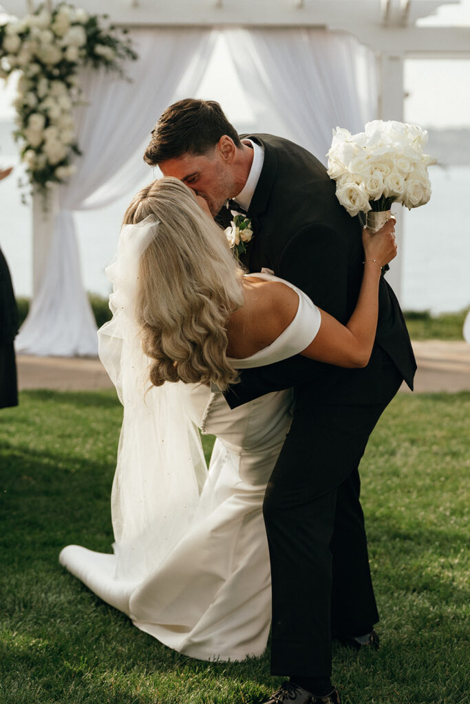 New England Beach Wedding | Aisha Lee Photography Cinematic Wedding Photographer | A couple in wedding attire shares a romantic kiss outdoors. The groom dips the bride, who holds a bouquet of white roses. Elegant white drapery and flowers adorn the background.