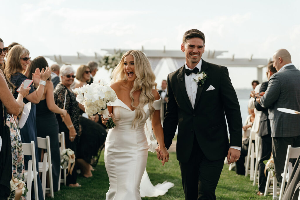 New England Beach Wedding | Aisha Lee Photography Cinematic Wedding Photographer | A joyous bride and groom walk hand in hand outdoors, the bride in an off-shoulder white gown holding white roses, smiling under a bright sky.