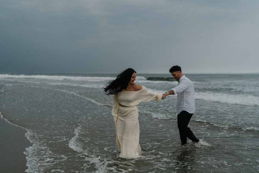 Oceanfront Weddings New England | Aisha Lee Photography Cinematic Wedding Photographer | A woman in a flowing cream dress smiles as she walks through shallow ocean waves, holding hands with a man in a white shirt. The sky is overcast.