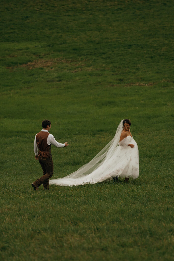 Cinematic Wedding Photographer Rhode Island | Aisha Lee Photography Cinematic Wedding Photographer | Bride in white gown with flowing veil smiles at groom in brown vest as they walk on a lush green field