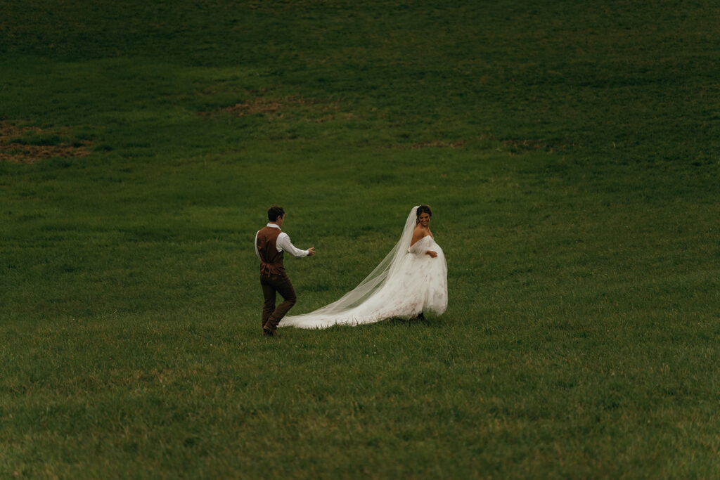 New England Wedding Venues | Aisha Lee Photography Cinematic Wedding Photographer | Bride in a flowing white gown and veil walks on a lush green field, smiling. A man in a vest and shirt follows. The scene feels joyful and serene.