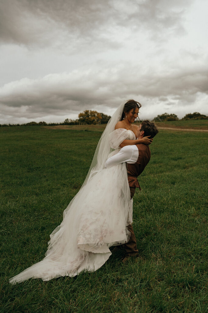 Cinematic Wedding Photographer Rhode Island | Aisha Lee Photography Cinematic Wedding Photographer | A groom lifts a bride in a flowing white dress and veil on a lush green field under an overcast sky