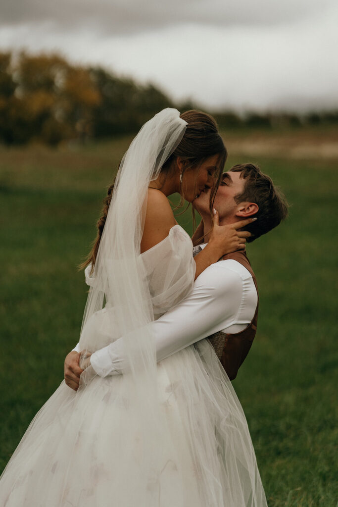 New England Wedding Venues | Aisha Lee Photography Cinematic Wedding Photographer | Bride in a flowing dress and veil is lovingly lifted by the groom, both smiling with closed eyes in a grassy field under a cloudy sky. Romantic and serene.