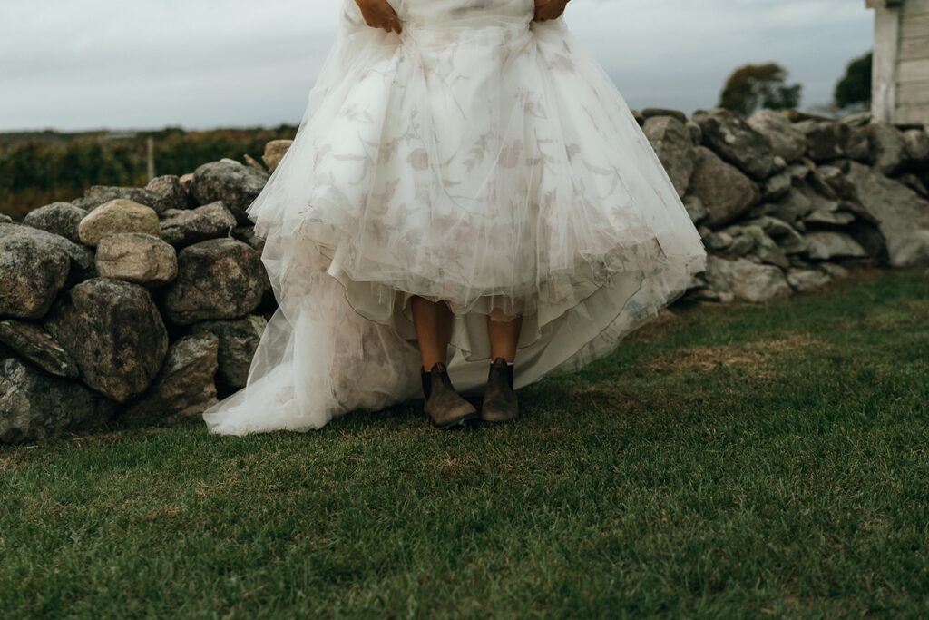 Close view of a floral wedding dress and boots standing on the grass, capturing a relaxed, fashion-forward bridal moment at beach wedding venues in New England.