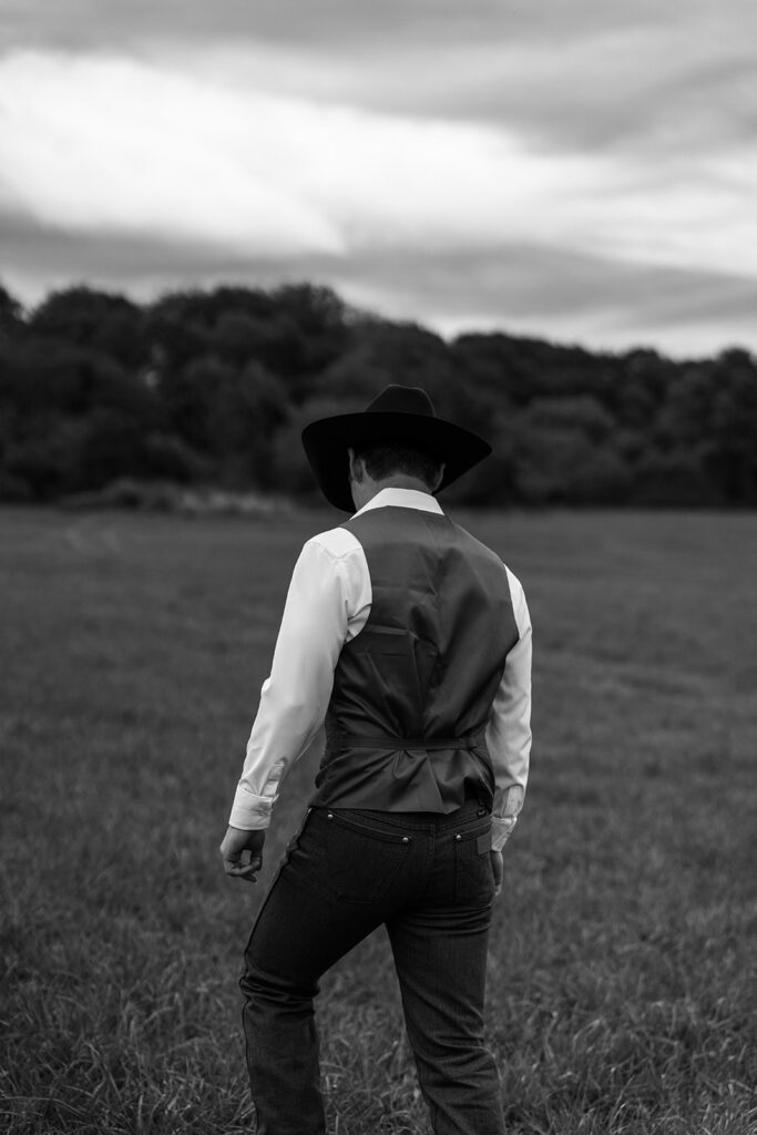 A groom in a vest and black hat walks alone through an open field under a dramatic sky, evoking the mood and character of destination coastal wedding venues in New England.