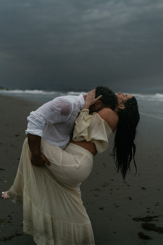 Traveling Cinematic Wedding and Elopement Photographer | Aisha Lee Photography Cinematic Wedding Photographer | A couple embraces passionately on a moody beach under stormy skies. The man in a wet white shirt lifts the woman in a flowing cream dress.