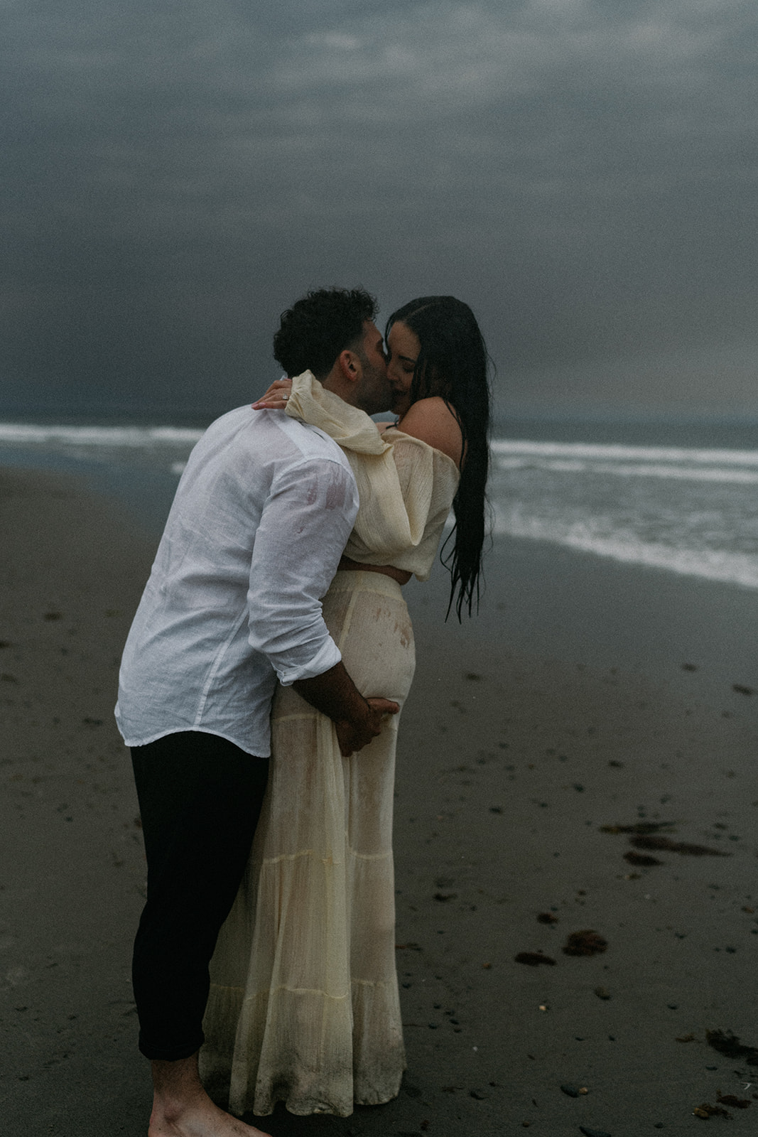 Destination Engagement Photographer New England | Aisha Lee Photography Cinematic Wedding Photographer | A couple embraces and kisses on a sandy beach under a moody, overcast sky.