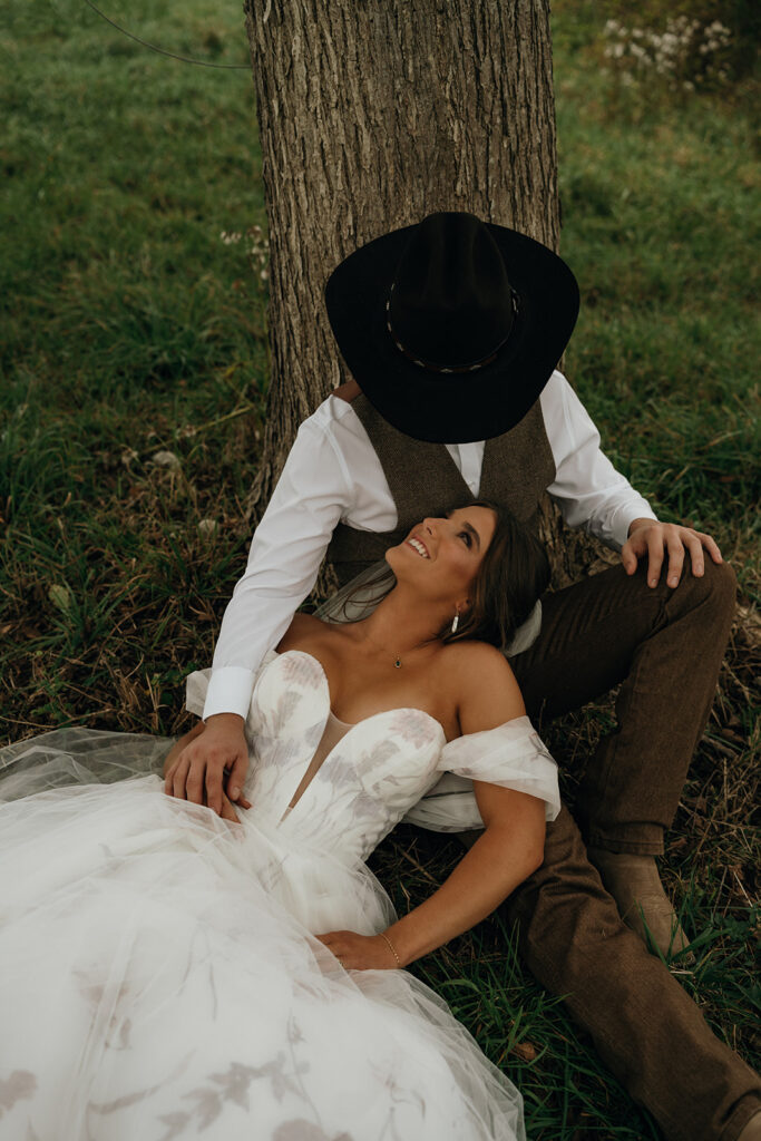 Unique Wedding Venues in New England | Aisha Lee Photography Cinematic Wedding Photographer | A bride in a white gown lies on green grass, smiling up at a groom in a cowboy hat and vest, leaning against a tree. The mood is romantic and serene.
