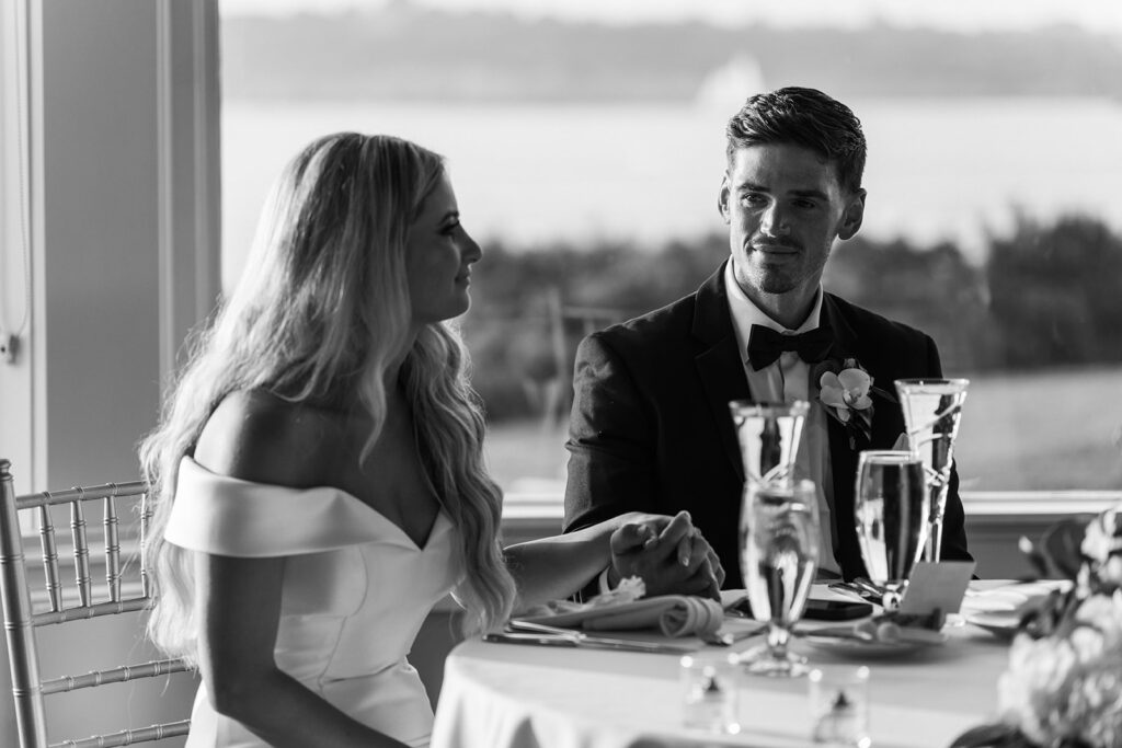 Coastal Wedding Venues New England | Aisha Lee Photography Cinematic Wedding Photographer | A couple sits at a table by a window overlooking a scenic view, holding hands. The bride is in a white dress, and the groom is in a tuxedo. The tone is intimate and serene.