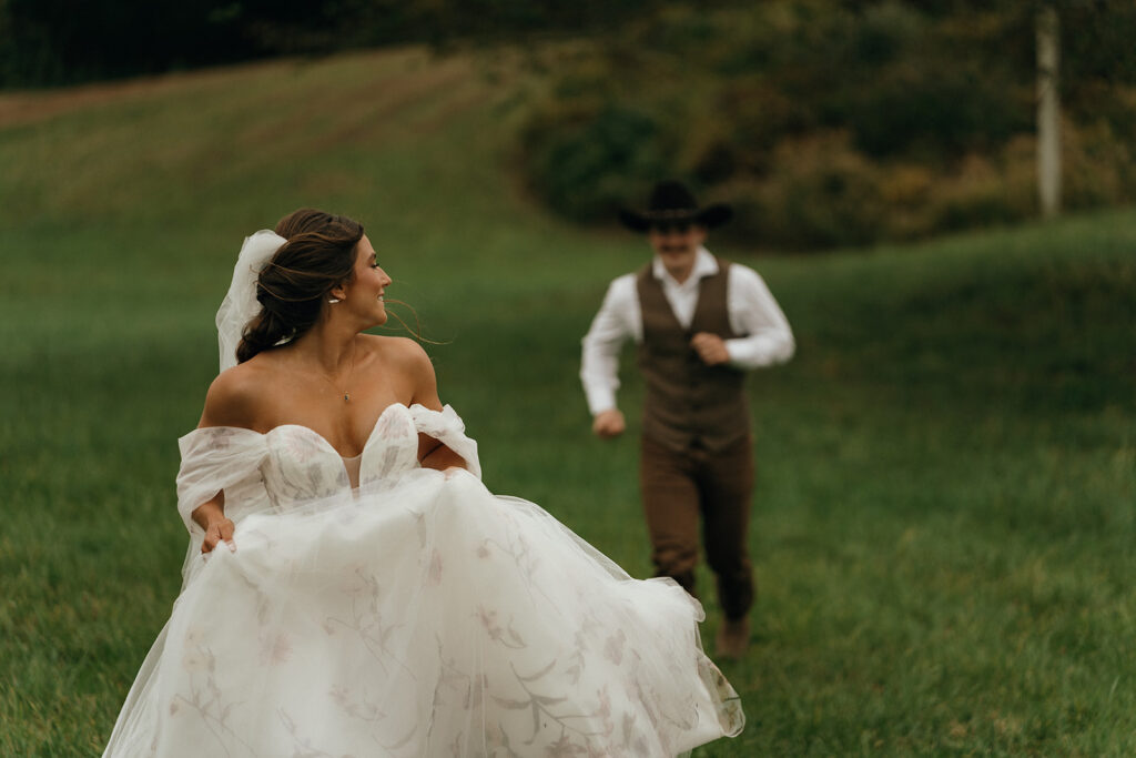 Unique Wedding Venues New England | Aisha Lee Photography Cinematic Wedding Photographer | A bride in an off-the-shoulder gown joyfully runs through a lush green field, looking back at a groom in Western attire with a cowboy hat.