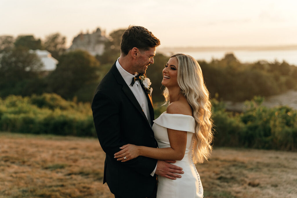 At sunset, the couple shares a quiet embrace overlooking rolling hills and water, highlighting the scenic beauty of coastal wedding venues in New England.