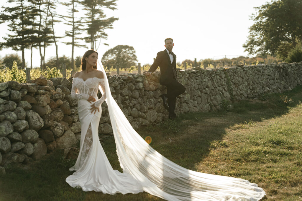 Bride and groom pose by a stone wall in golden light, with her long veil trailing across the grass for a dramatic finish. A fine-art portrait inspired by vineyard wedding venues New England.