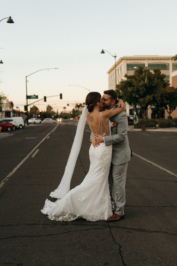 Destination Wedding and Elopement Photographer | Aisha Lee Photography Cinematic Wedding Photographer | A bride in a lace gown and veil kisses a groom in a gray suit on a quiet street at sunset.
