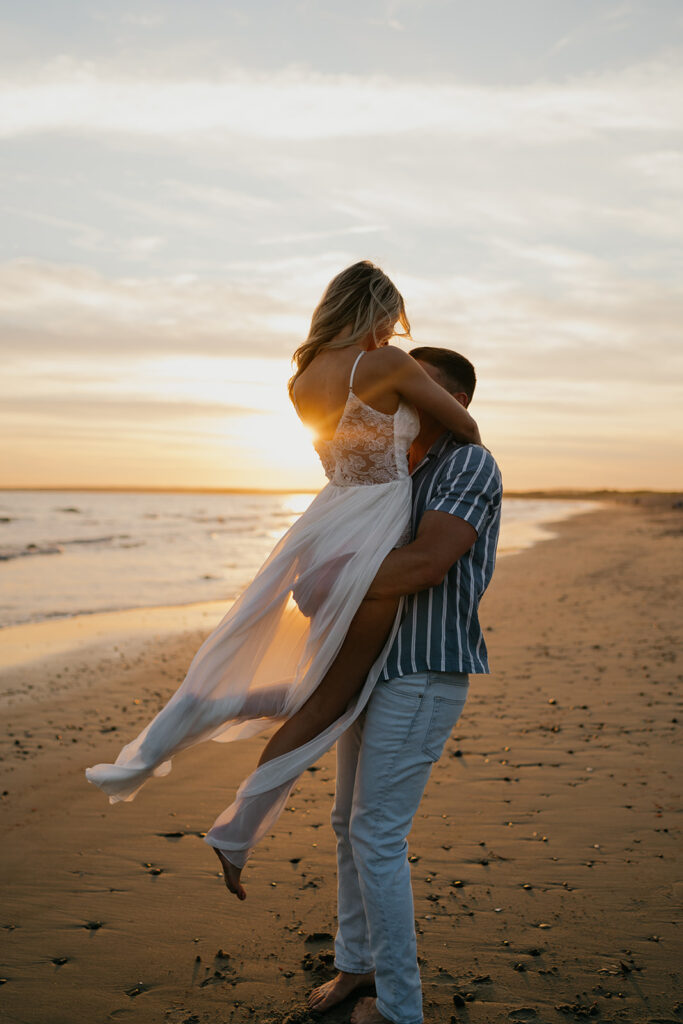 Oceanfront Weddings New England | Aisha Lee Photography Cinematic Wedding Photographer | A couple joyfully embraces on a beach at sunset. The woman, in a flowing white dress, is lifted by the man, creating a romantic and serene scene.