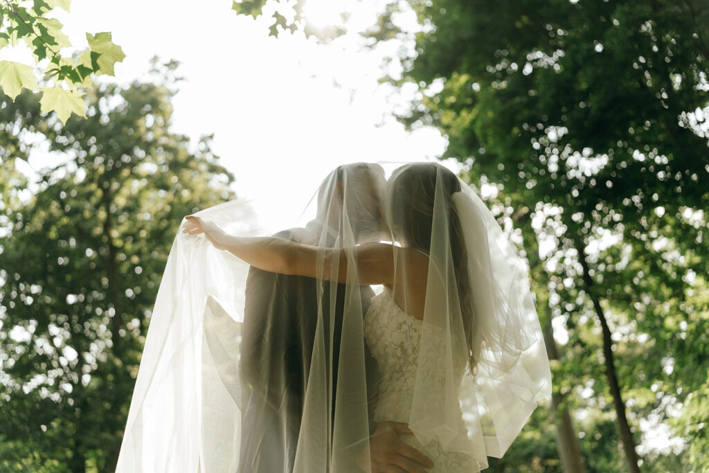 Unique Wedding Venues in New England | Aisha Lee Photography Cinematic Wedding Photographer | A bride and groom share a kiss under a veil in a sunlit, wooded setting. The mood is romantic and intimate, with dappled sunlight filtering through trees.