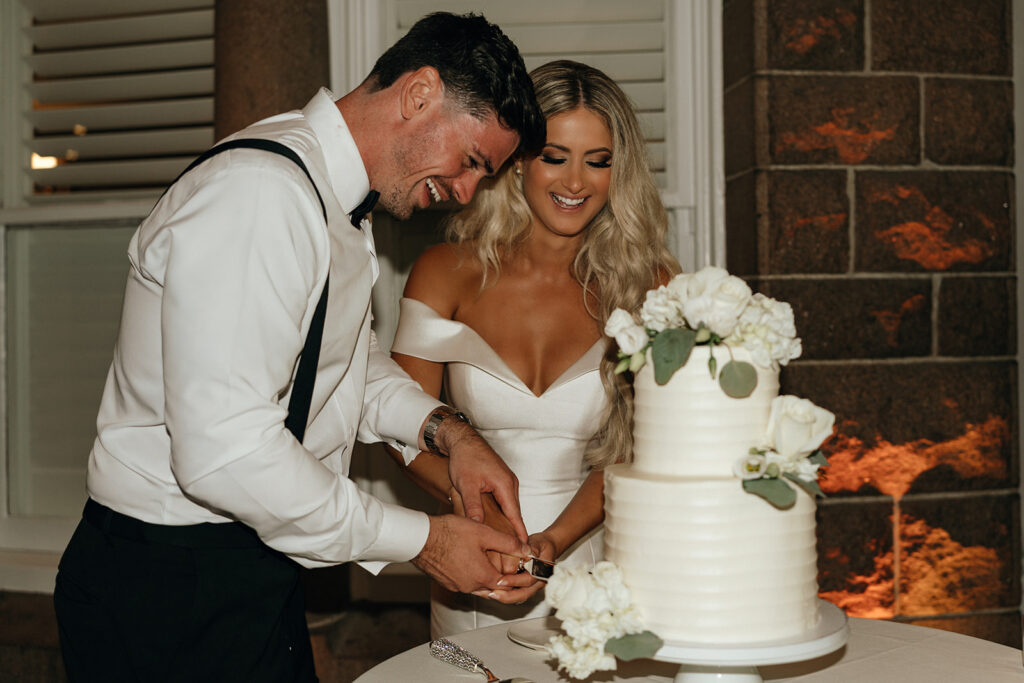 The couple laughs together while cutting their wedding cake, surrounded by warm candlelight and joyful energy that defines luxury wedding venues in New England.
