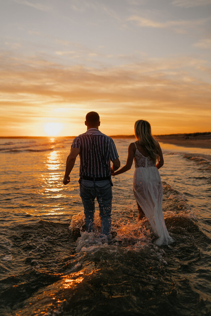 Oceanfront Weddings New England | Aisha Lee Photography Cinematic Wedding Photographer | A couple holds hands while walking through shallow waves at sunset, casting long shadows on the water. The sky is a warm mix of orange and gold, creating a romantic atmosphere.