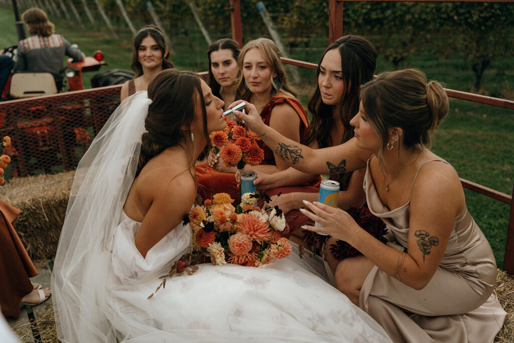 Unique Wedding Venues New England | Aisha Lee Photography Cinematic Wedding Photographer | A bride in a white gown holds a colorful bouquet. A friend applies lipstick while others watch, holding drinks. The setting is outdoors, with a joyful vibe.