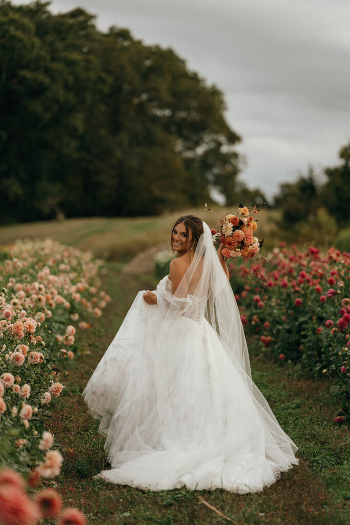 Castle Wedding Venues in New England | Aisha Lee Photography Cinematic Wedding Photographer | A bride in a flowing white dress and veil smiles while holding a bouquet of orange flowers. She stands on a path lined with blooming dahlias.