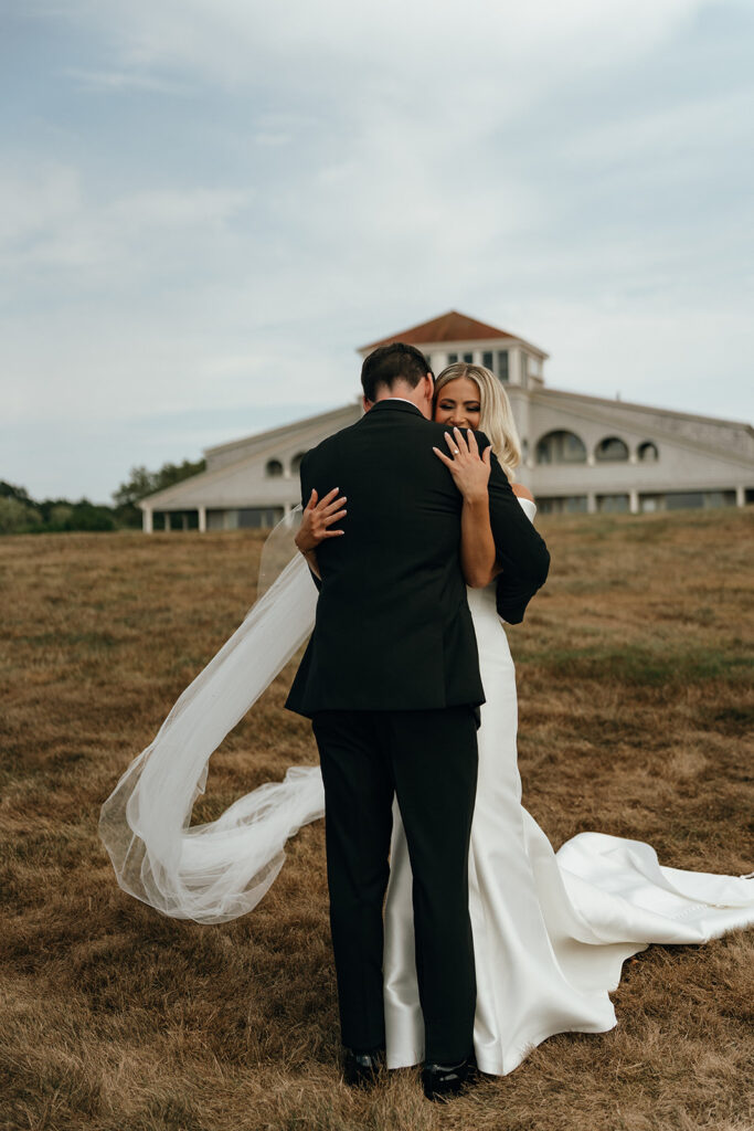 Unique Wedding Venues in New England | Aisha Lee Photography Cinematic Wedding Photographer | A bride and groom embrace on a grassy field, with the bride's flowing veil trailing behind. A large building is visible in the background under a cloudy sky.