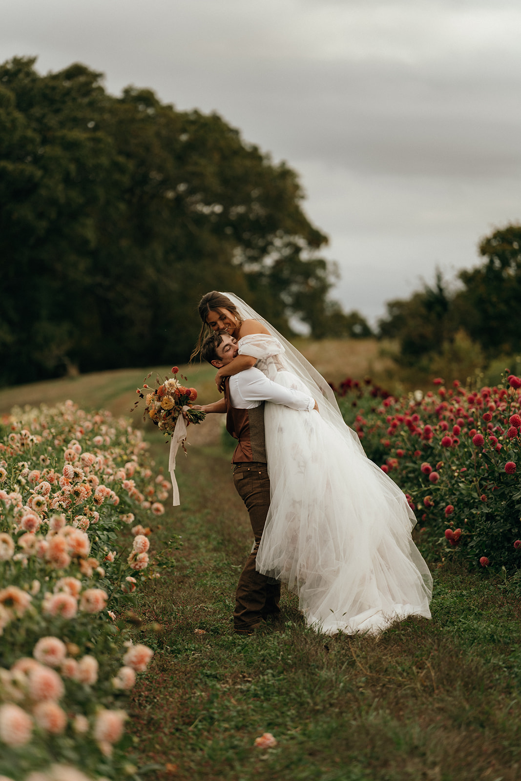 Moody Wedding Photographer Rhode Island | Aisha Lee Photography Cinematic Wedding Photographer | A groom playfully lifts the bride in a vibrant flower field. She smiles, holding a bouquet. Overcast sky and lush greenery