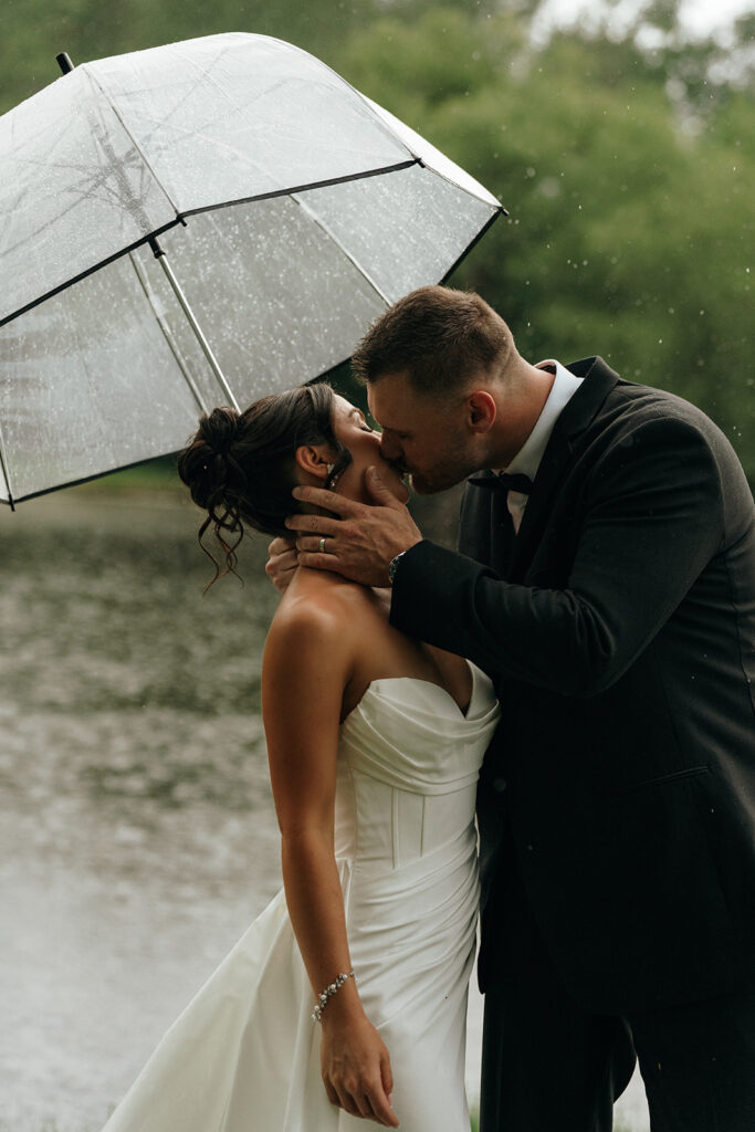 Oceanfront Weddings New England | Aisha Lee Photography Cinematic Wedding Photographer | A bride and groom share a romantic kiss under a clear umbrella in the rain by a lakeside. The bride wears a strapless white gown, and the groom is in a black suit.