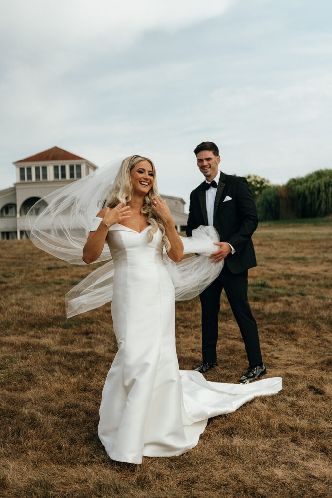 Unique Wedding Venues in New England | Aisha Lee Photography Cinematic Wedding Photographer | Bride in a white gown and veil laughs joyfully while standing with a groom in a black tuxedo on a grassy field. Elegant building visible in the background.