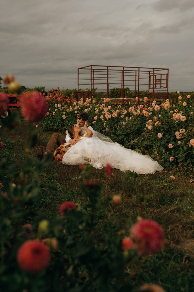 Unique Wedding Venues in RI | Aisha Lee Photography Cinematic Wedding Photographer | Bride and groom sit romantically in a lush flower field. They are surrounded by pink blooms under a cloudy sky, exuding tranquility and love.