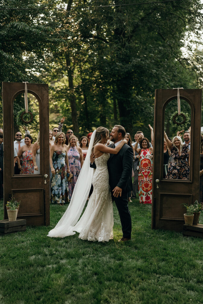 Unique Wedding Venues in New England | Aisha Lee Photography Cinematic Wedding Photographer | Bride and groom share a kiss outdoors, framed by rustic doors. Guests clap and smile behind them, surrounded by lush trees, creating a joyful atmosphere.