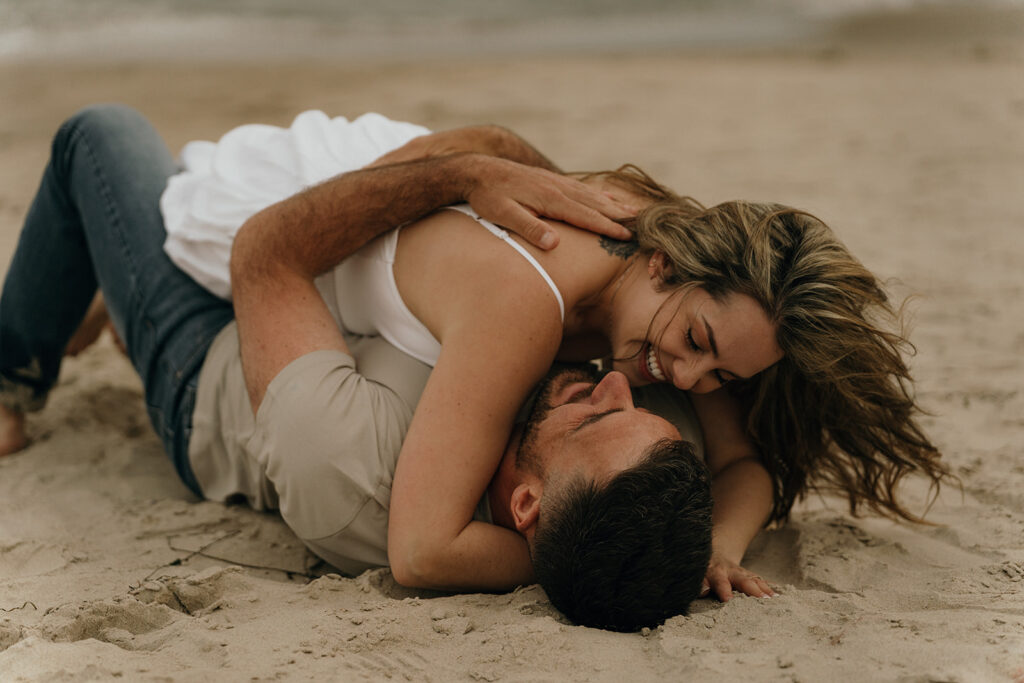 The couple laughs together while lying on the sand, a playful and intimate portrait that fits beautifully with Rhode Island beach wedding venues.