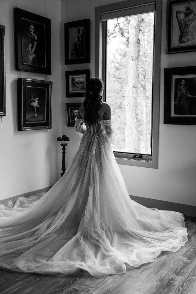 Elopement Packages in RI | Aisha Lee Photography Cinematic Wedding Photographer | A bride in an elegant gown stands near a window in an art-filled room, gazing outside. The mood is contemplative, with soft lighting highlighting the dress's details.