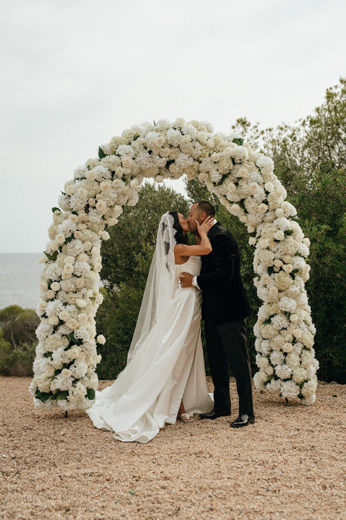 Unique Wedding Venues in New England | Aisha Lee Photography Cinematic Wedding Photographer | A bride and groom share a kiss under a large white floral arch outdoors. The bride wears a flowing gown and veil; the groom is in a black suit. Romantic and serene atmosphere.