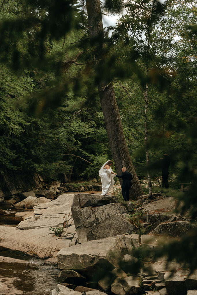 Unique Wedding Venues in New England | Aisha Lee Photography Cinematic Wedding Photographer | Wedding couple stands on rocky terrain by a stream, surrounded by lush green trees. The setting is serene and natural, conveying a peaceful, romantic mood.