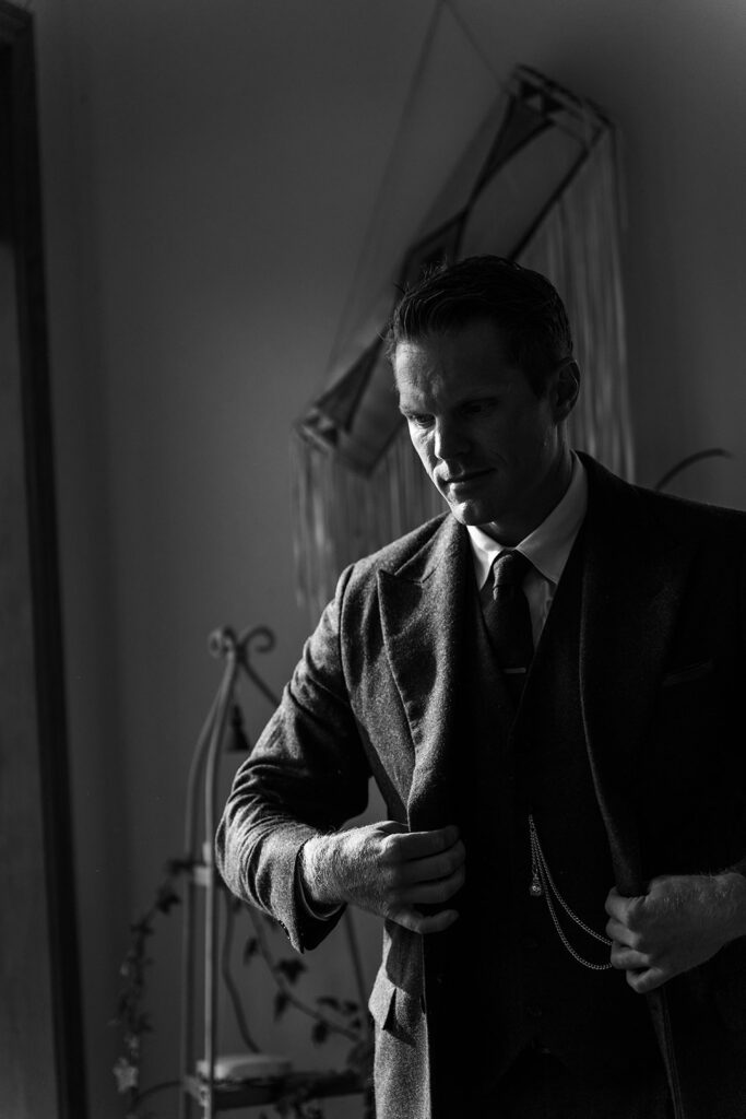 Top New England Wedding Venues | Aisha Lee Photography Cinematic Wedding Photographer | A man in a suit adjusts his jacket in a dimly lit room, conveying a somber, contemplative mood. Shadows accentuate his features and the vintage decor.