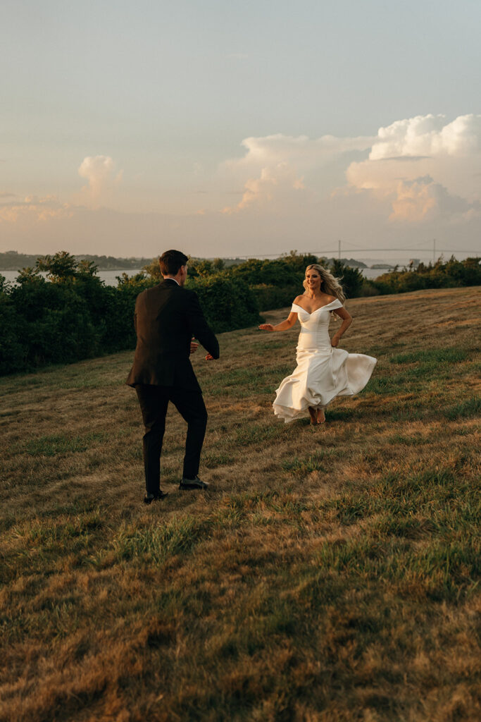 Oceanfront Weddings New England | Aisha Lee Photography Cinematic Wedding Photographer | A bride in a white gown joyfully runs towards a groom in a suit on a sunlit hill. The background features trees, a distant bridge, and cloudy sky.