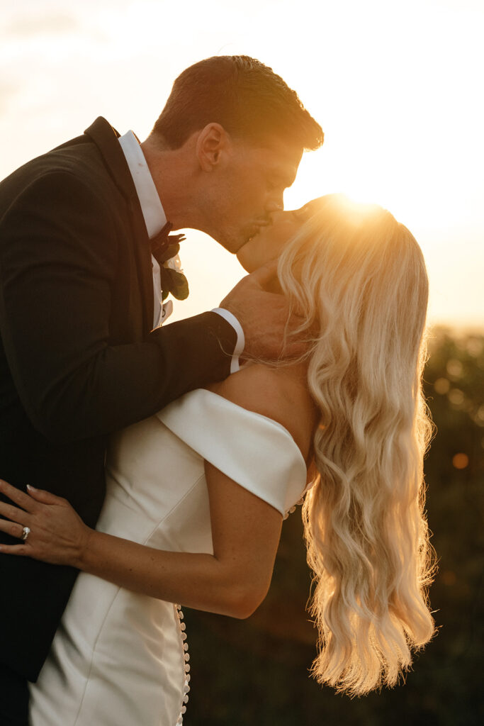New England Wedding Venues | Aisha Lee Photography Cinematic Wedding Photographer | A couple kisses at sunset, the man in a black suit gently holding a woman in a white off-shoulder dress. Her long blonde hair shines in the warm light.
