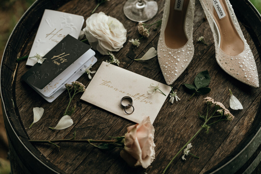 Styled flat lay of wedding details including rings, invitation, and embellished heels arranged on a rustic table, complementing the refined aesthetic of a Castle New England celebration.
