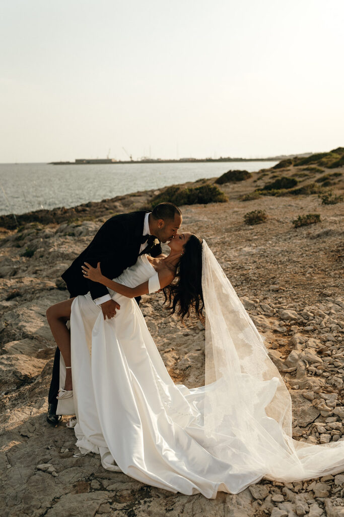 Oceanfront Weddings New England | Aisha Lee Photography Cinematic Wedding Photographer | A couple shares a romantic kiss by the seaside at sunset. The bride's long veil flows over rocky terrain, adding elegance to the serene, intimate moment.