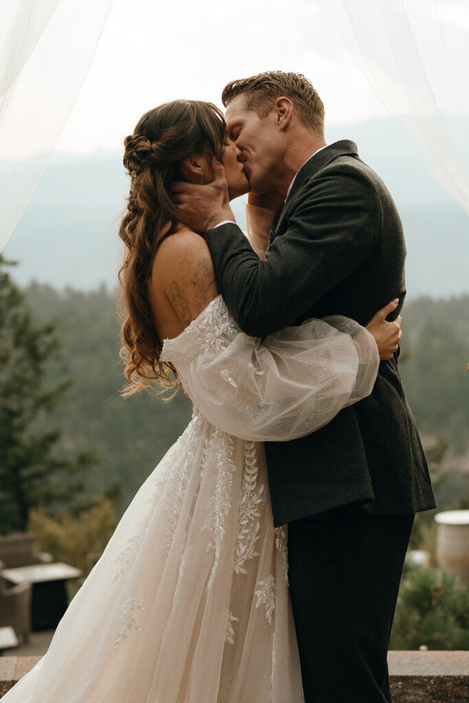 Oceanfront Weddings New England | Aisha Lee Photography Cinematic Wedding Photographer | A bride and groom share a romantic kiss outdoors, framed by sheer white drapes. The bride wears a lace gown, and the groom is in a dark suit, against a forested mountain backdrop.