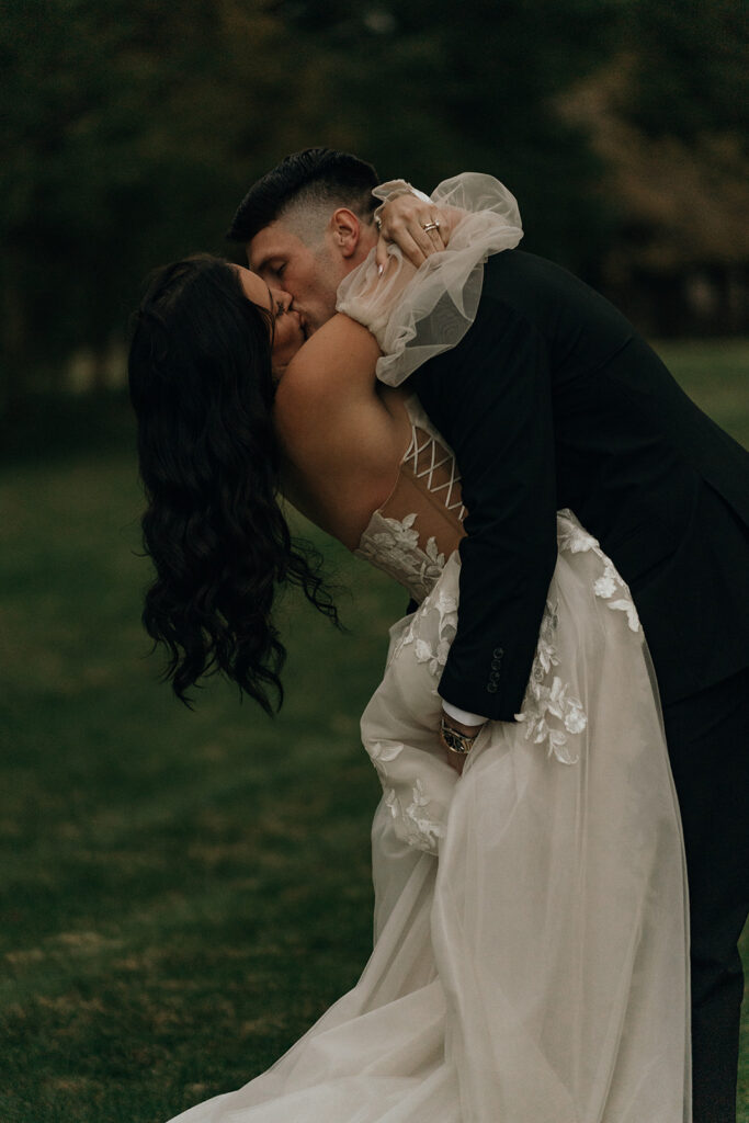 Bride and groom share a kiss on a grassy lawn at dusk, framed by deep green tones and soft movement in the dress. A romantic moment at a vineyard wedding venue in New England.